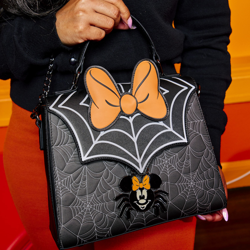 Person holding the Minnie Mouse Spider Crossbody Bag against an orange background. The bag features a big spiderweb on the front flap, with orange Minnie Mouse ears and Minnie Mouse as a spider on the front.